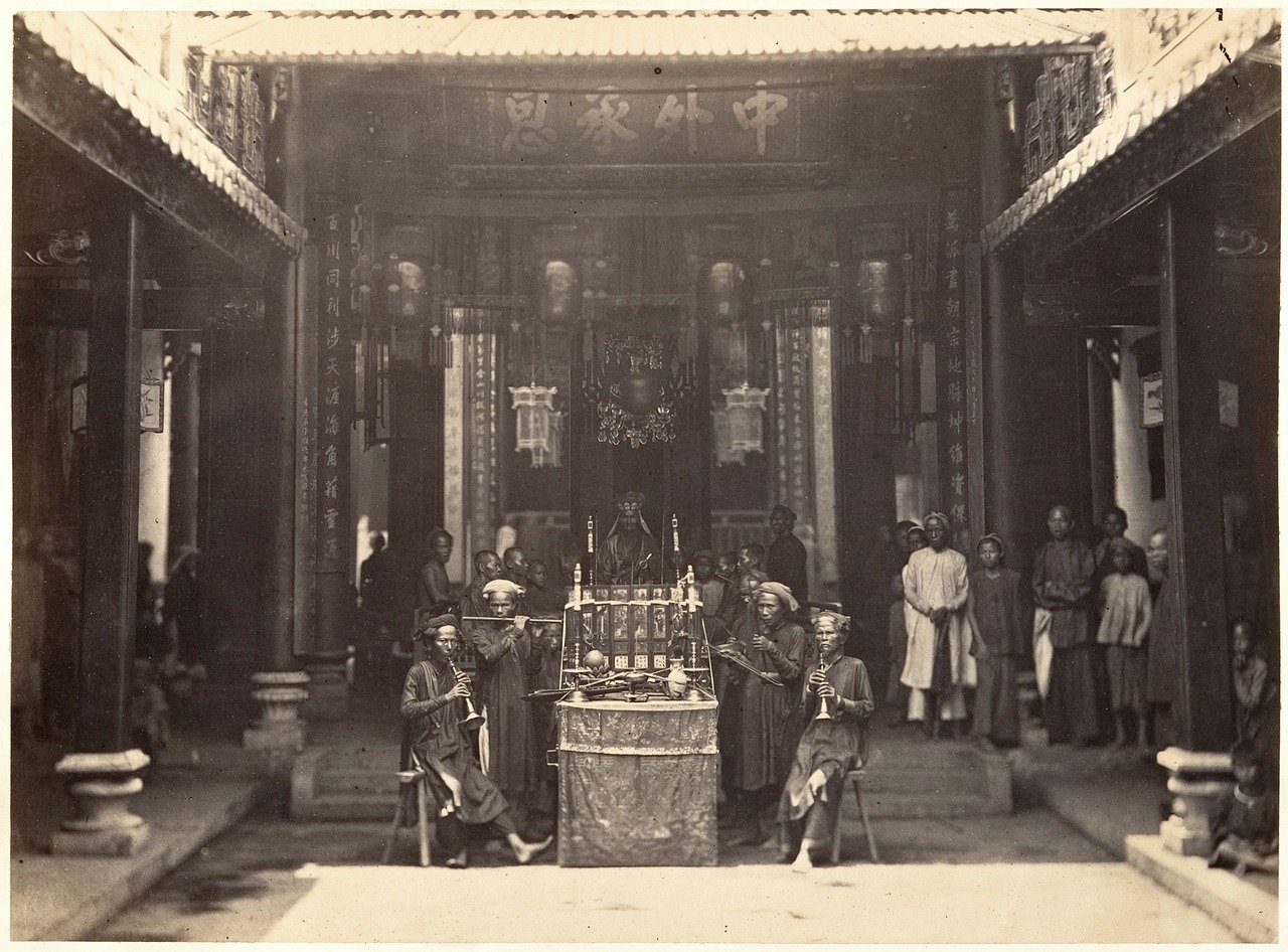 Vintage black and white photo of traditional Chinese musicians playing instruments in a temple courtyard with onlookers