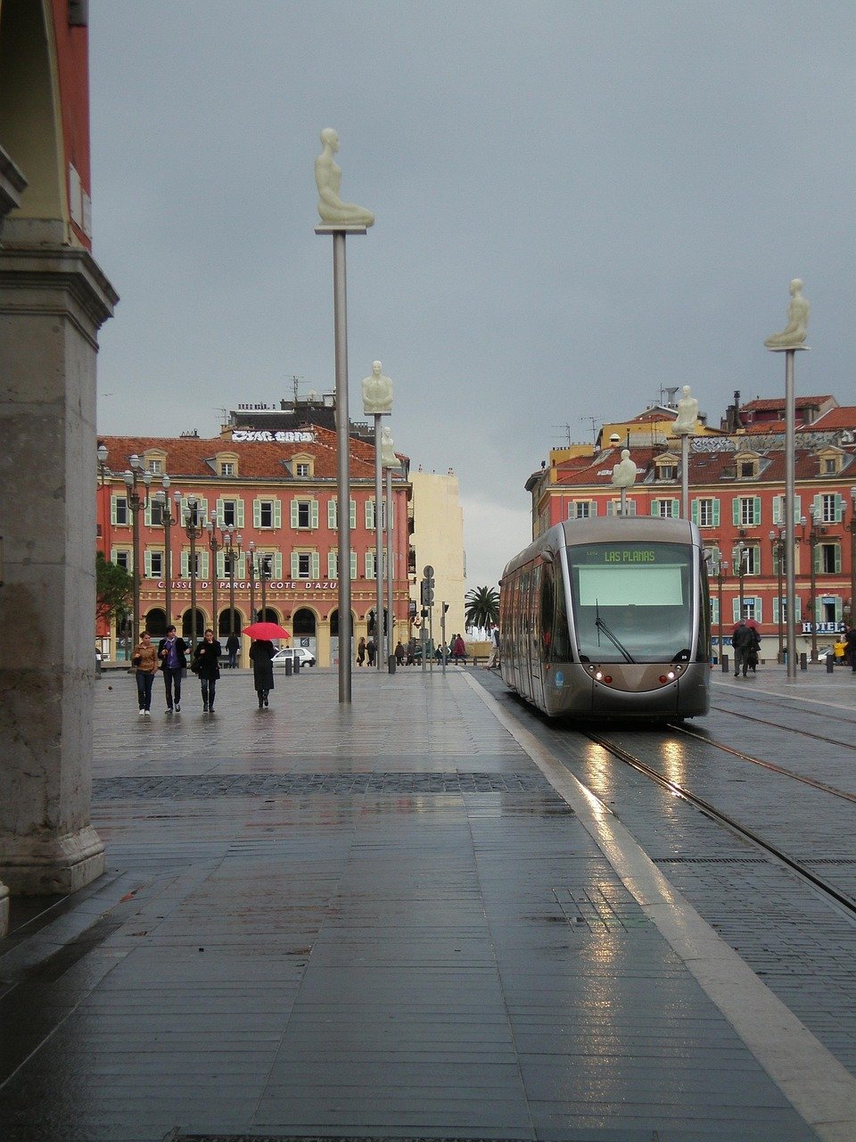 Modern tram on wet street with unique seated human sculptures on poles in a European city square