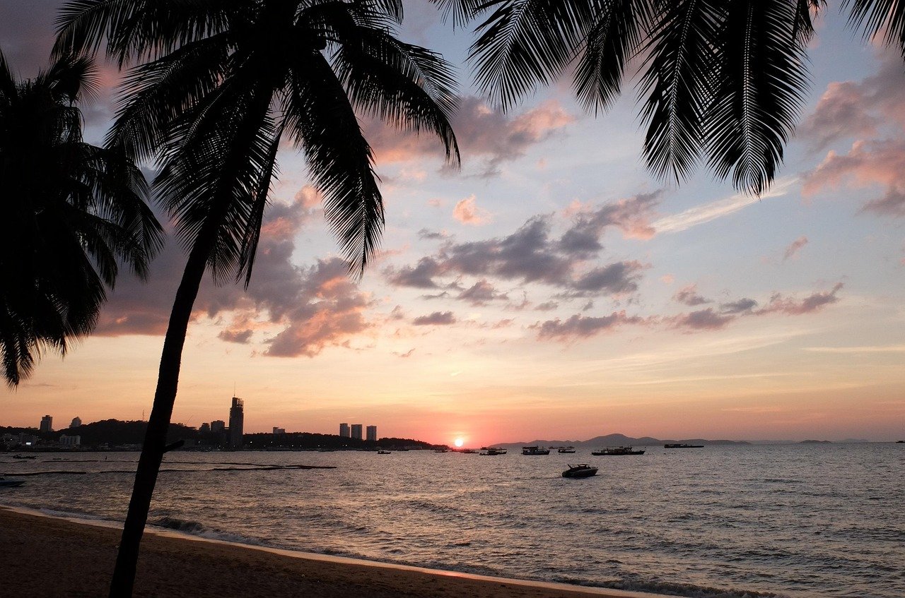 Sunset over a tropical beach with palm trees, calm sea, boats, and distant city skyline under colorful sky