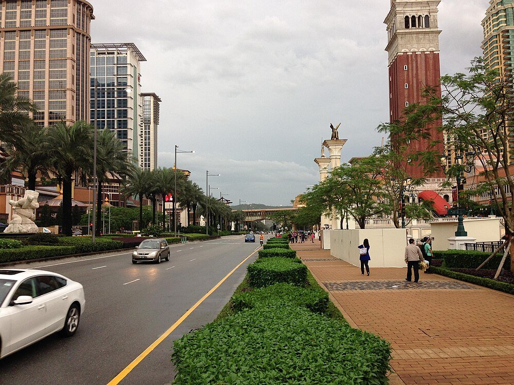 City street with buildings and pedestrians Urban street with modern buildings, palm trees, cars, and pedestrians under a cloudy sky in a cityscape setting
