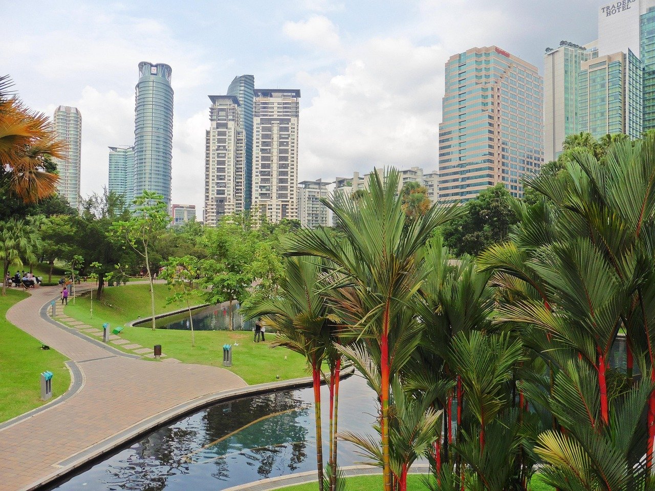 Urban park with walking path, pond, palm trees, and modern high-rise buildings in the background under a cloudy sky