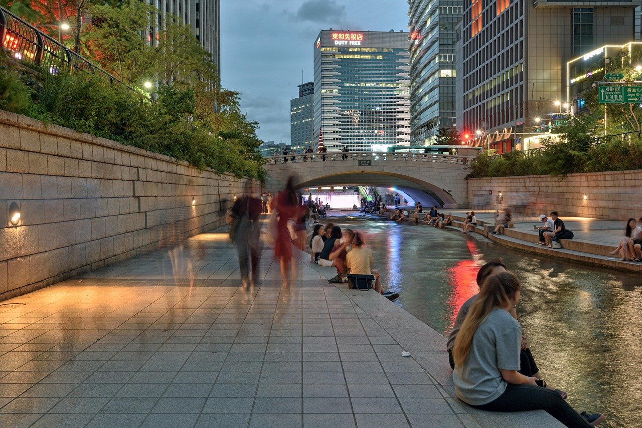 Evening urban scene with people relaxing by a river in a city park surrounded by modern buildings and illuminated bridge
