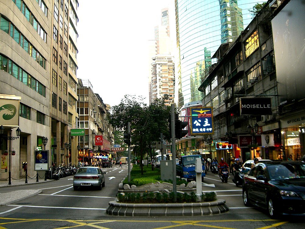 Urban street scene with vehicles and shops Busy urban street with cars, motorcycles, and shops in a city with modern and older buildings, featuring signs in Chinese and English.