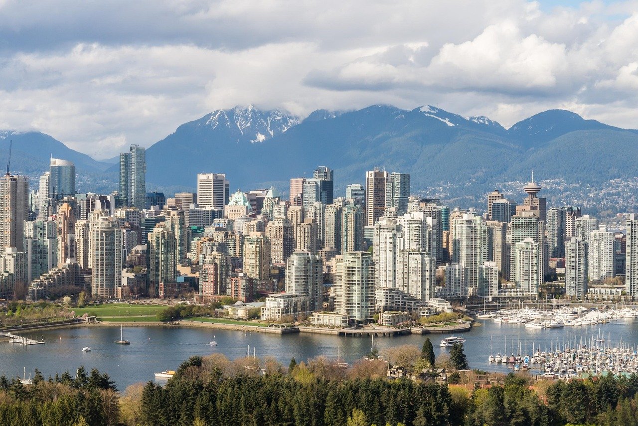 Vancouver city skyline with modern high-rise buildings, waterfront, and mountains in the background under cloudy sky