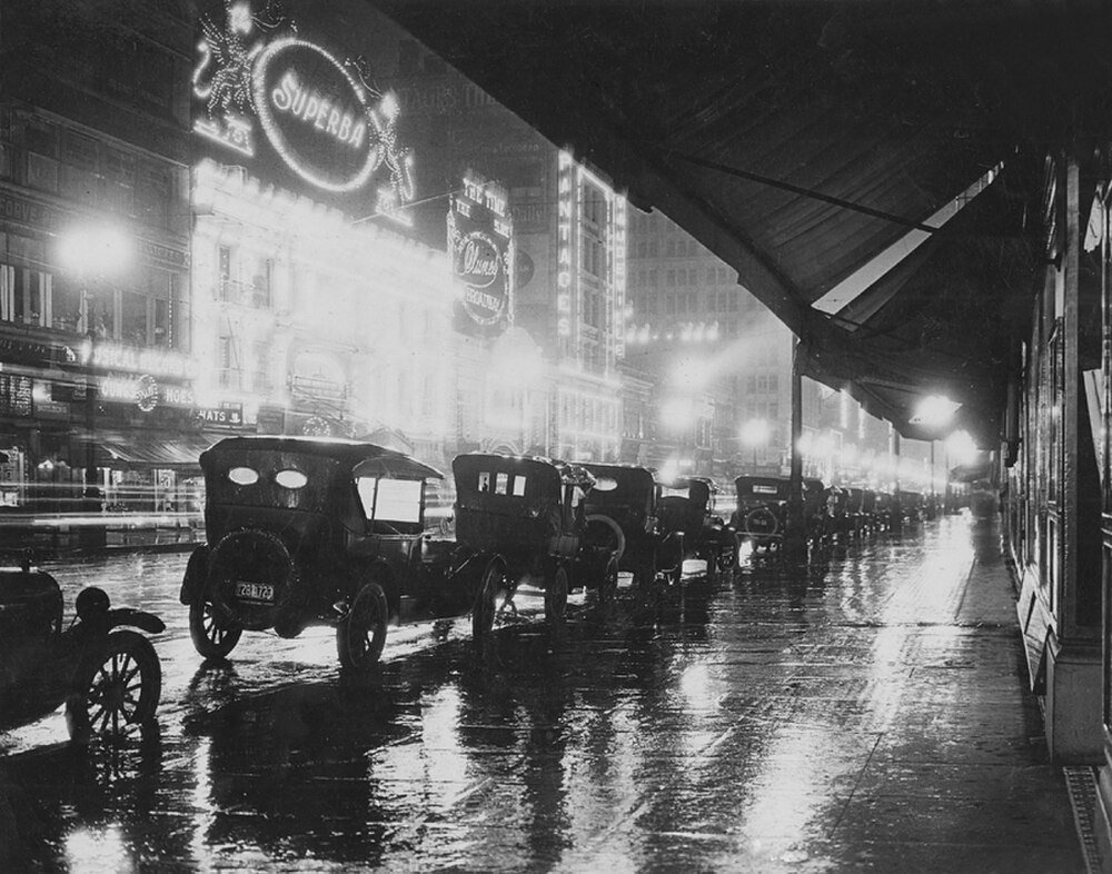 Vintage black and white photo of early 20th century cars parked on a rainy city street with bright neon theater signs