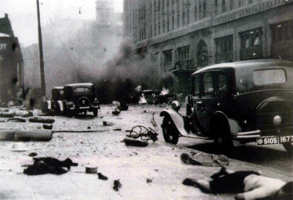Black and white photo of a chaotic street scene with vintage cars, smoke, debris, and fallen bodies during a historical conflict.
