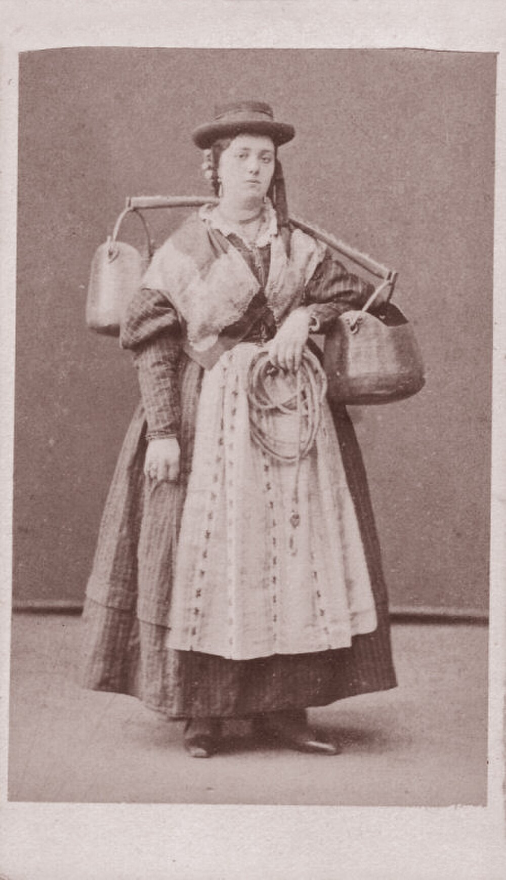 Vintage photo of woman with yoke and containers Vintage sepia photo of a woman in traditional attire carrying two large containers with a yoke