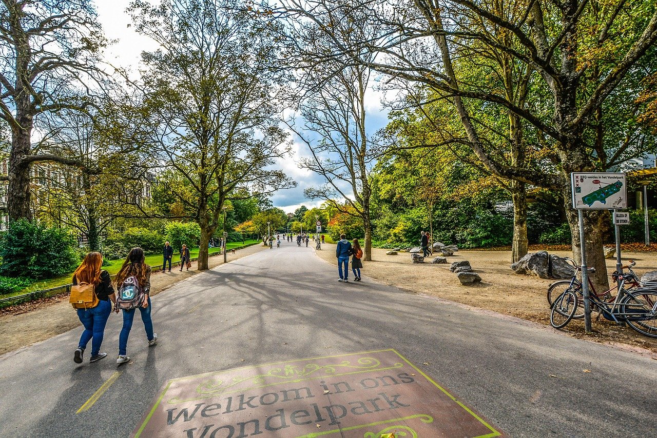 People walking and cycling in Vondelpark Amsterdam with trees and signage welcoming visitors to the park