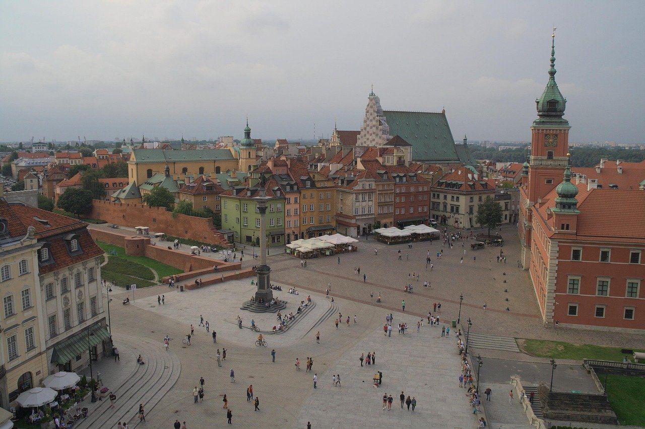 Aerial view of Warsaw Old Town Market Square with colorful historic buildings, Sigismund's Column, and tourists walking around.