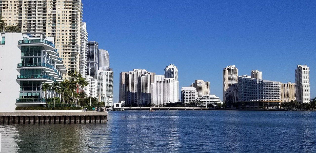 Waterfront city skyline with modern high-rise buildings under clear blue sky, reflecting on calm water surface