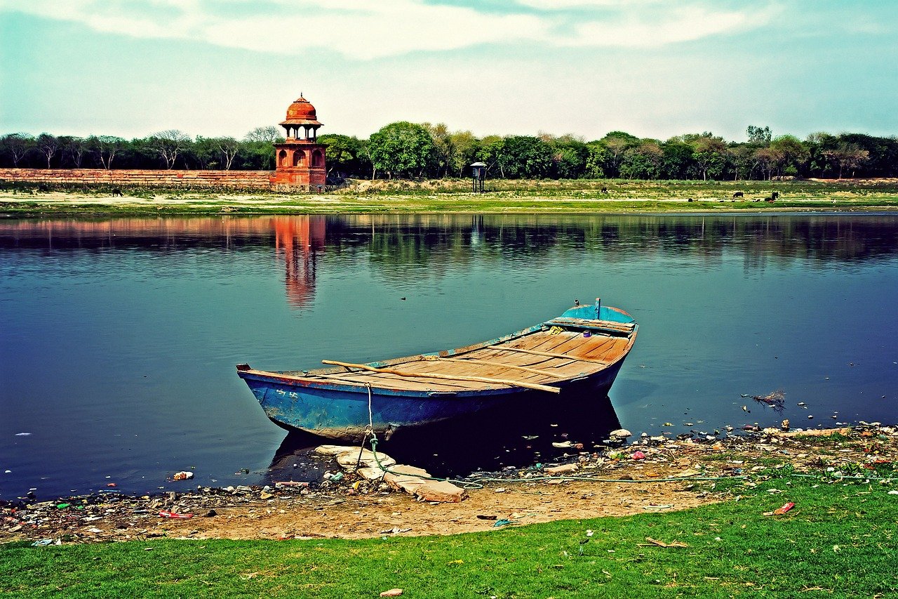 Wooden boat tied on riverbank with historic red sandstone pavilion and green trees reflected in calm water