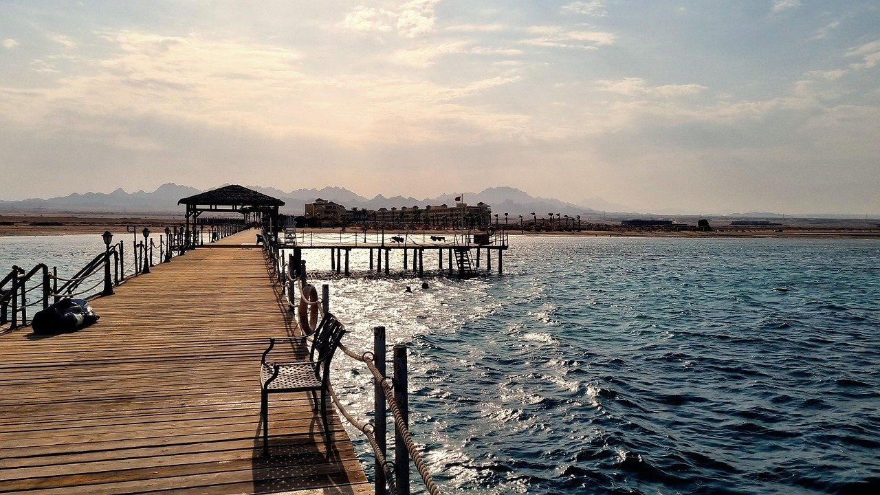 Wooden pier extending over blue ocean water with distant mountains and cloudy sky at sunset
