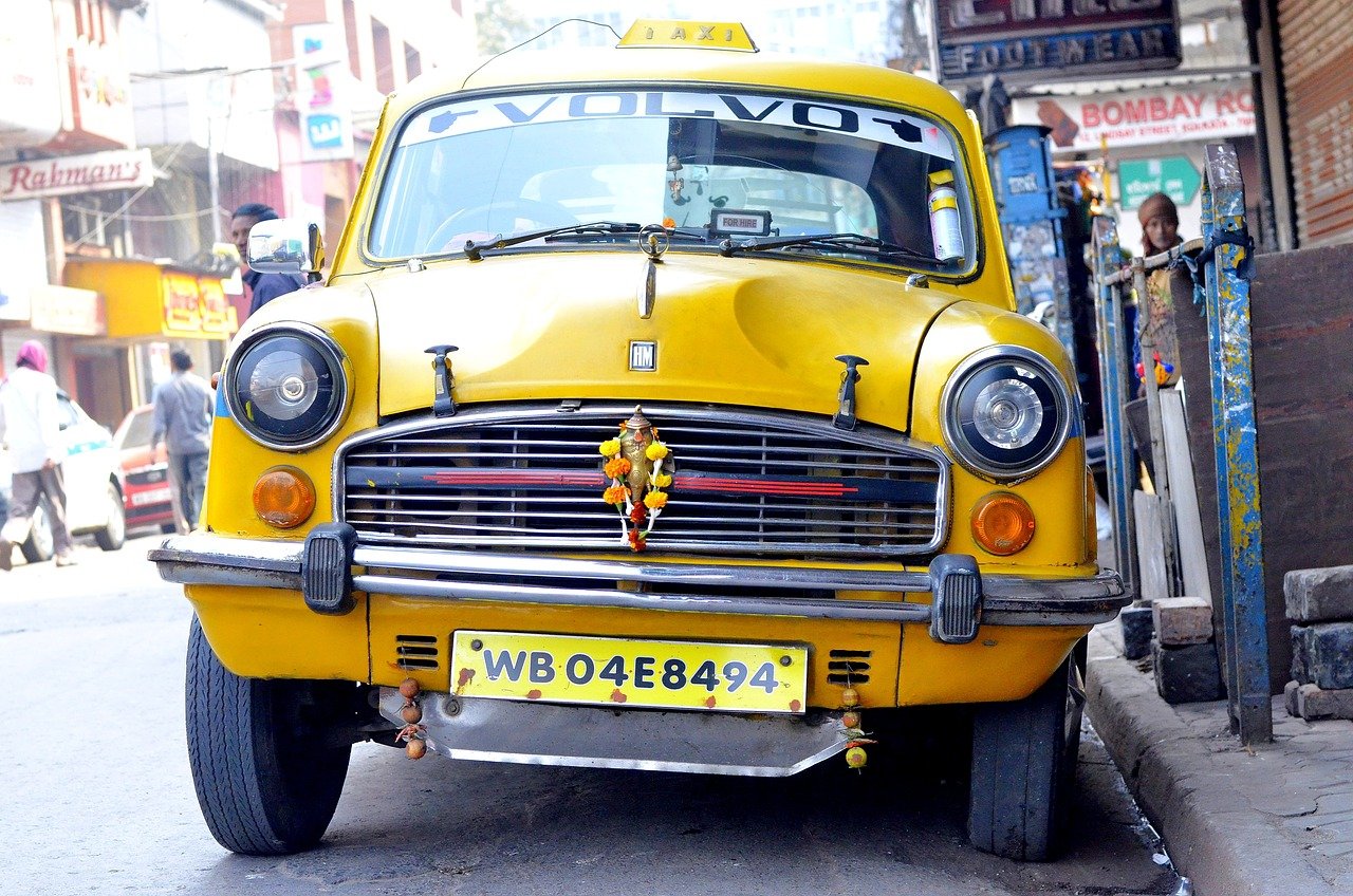 Yellow vintage taxi car with WB04E8494 license plate parked on street in busy urban area