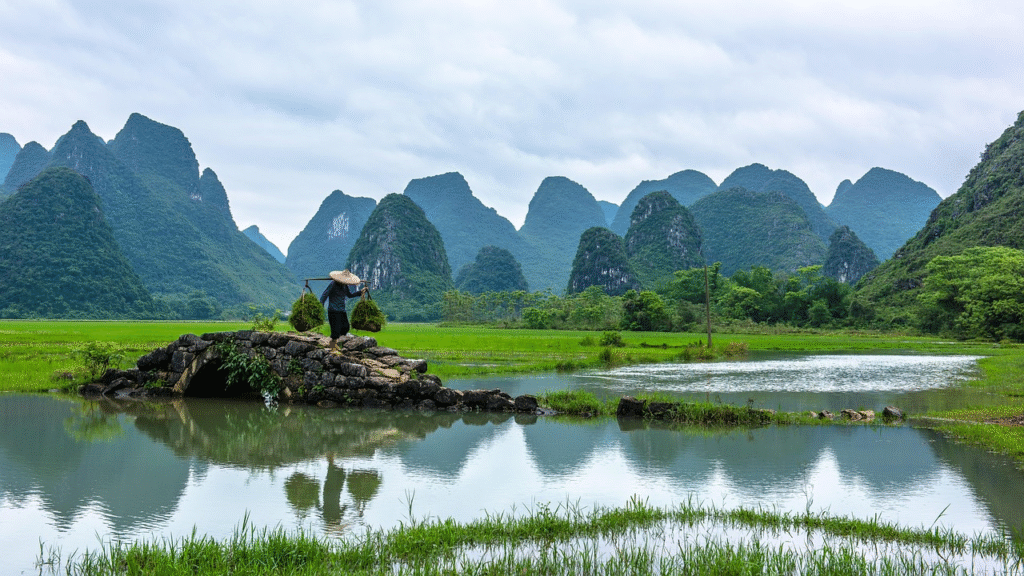 A farmer carries baskets across a stone bridge in the Guilin countryside — the iconic karst-mountain limestone peaks rising in the background, the landscape that appears on the back of the Chinese 20 yuan banknote