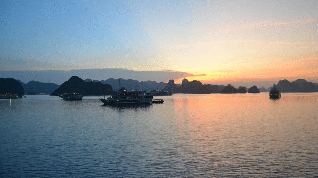 Sunset over Ha Long Bay — traditional Vietnamese junk boats anchored among the iconic limestone karst islands rising from emerald water