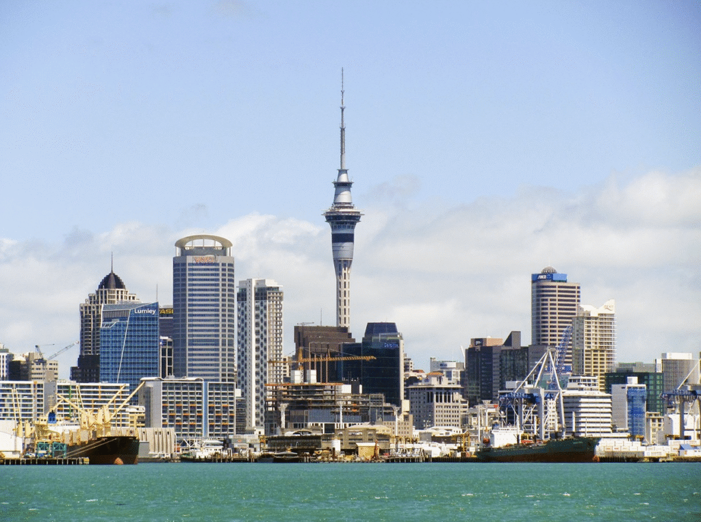 Auckland city skyline featuring the iconic Sky Tower, the waterfront harbor, and cargo ships — New Zealand's largest city framed against the Pacific Ocean