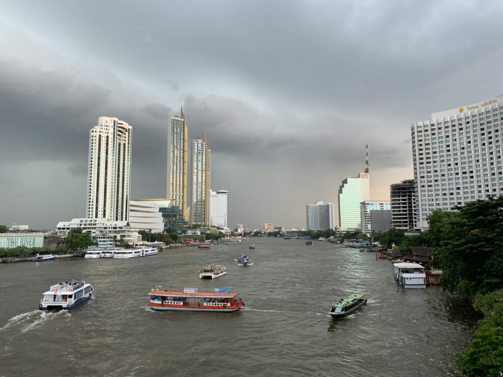 View of the Chao Phraya River through Bangkok with traditional boats and modern skyscrapers under cloudy sky
