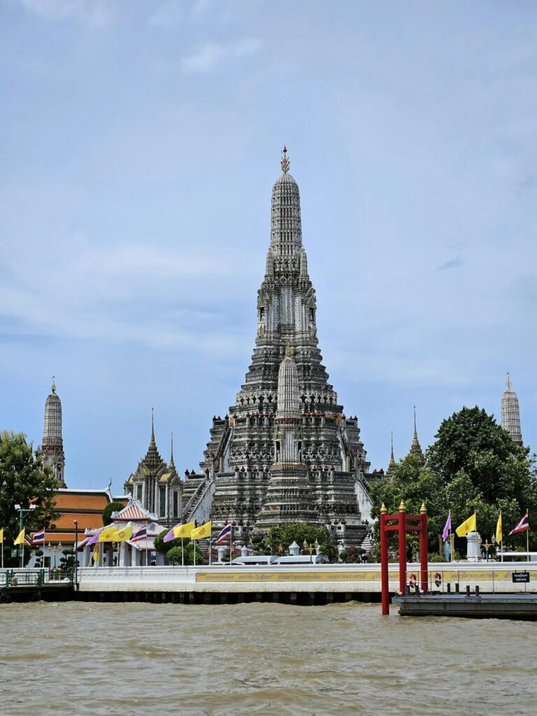 Wat Arun temple seen from across the Chao Phraya River in Bangkok, showcasing its 75-meter porcelain-covered prang under clear sky