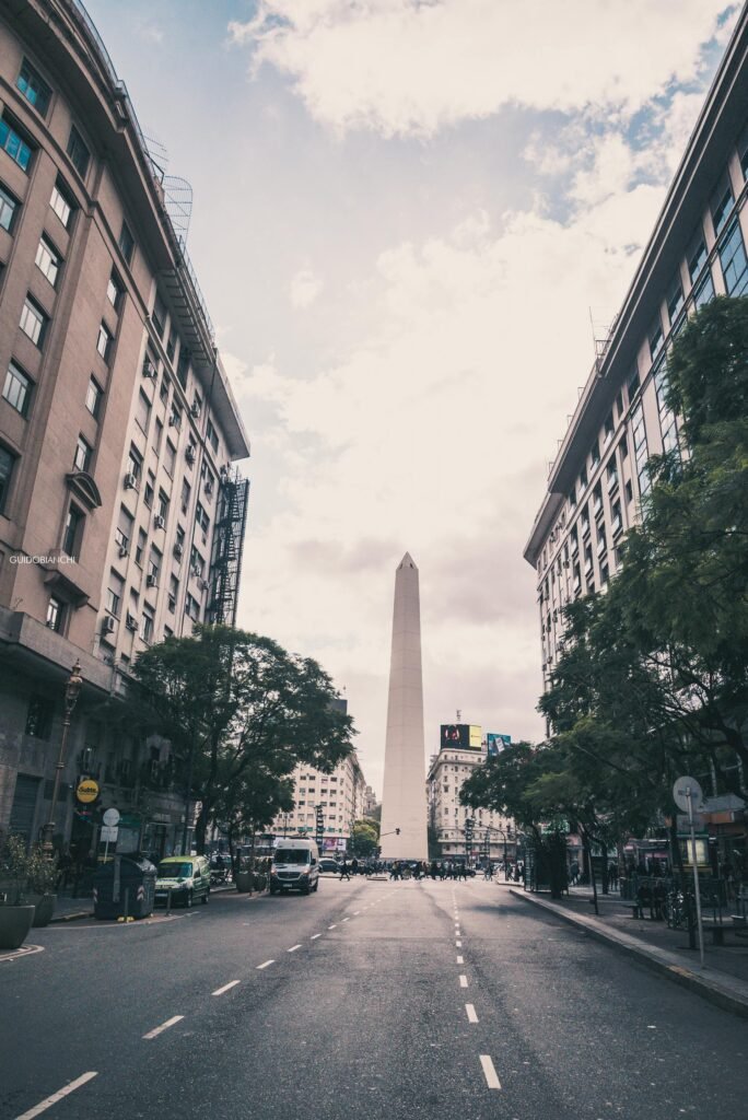 The iconic Obelisk of Buenos Aires towering between tall buildings on Avenida Corrientes — the 67-meter monument erected for the city's 400th anniversary in 1936, the symbolic geographic center of Argentina's capital