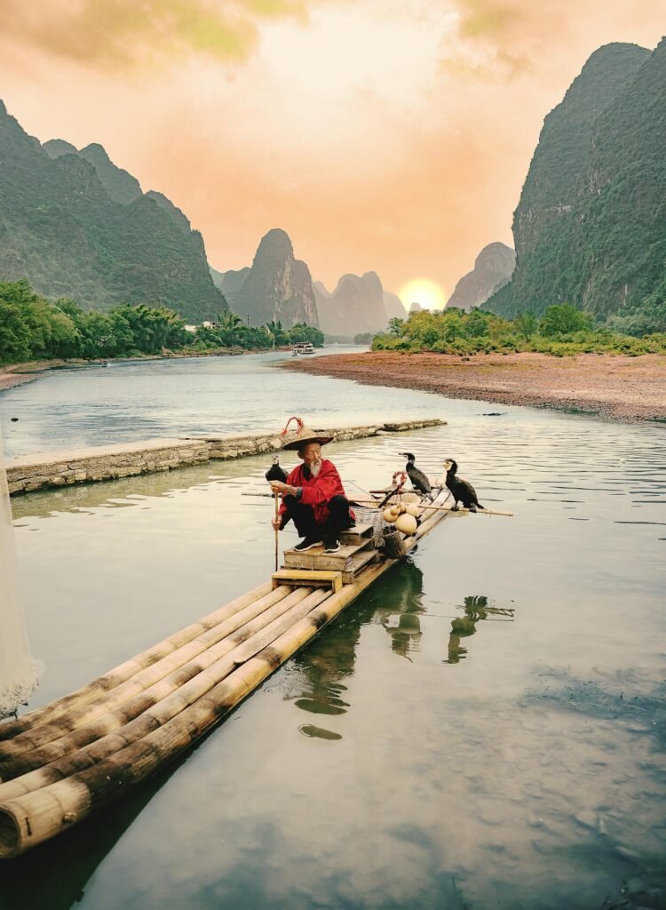 An elderly fisherman on a traditional bamboo raft fishing with trained cormorants on the Li River at sunset — the centuries-old practice that remains a Guilin cultural emblem