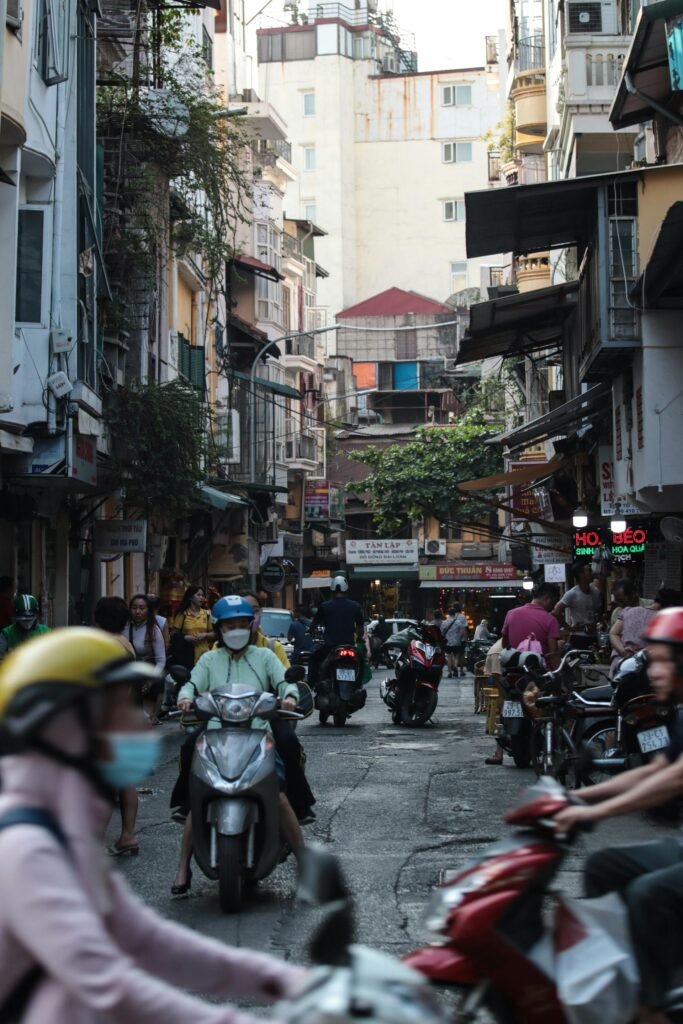 Lively street in Hanoi's Old Quarter — the medieval commercial heart of the city with narrow tube-house buildings, scooters, and pedestrians weaving through the 36 historic guild streets