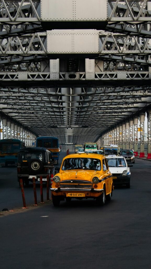 Iconic yellow Hindustan Ambassador taxi beneath the Howrah Bridge in Kolkata — the city's signature transit fleet capturing the working morning urban life along the Hooghly River