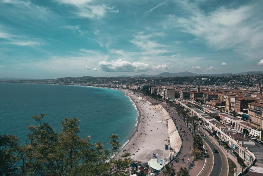 Stunning aerial view of Nice's beachfront under a clear summer sky — the UNESCO-listed Promenade des Anglais curving along the Baie des Anges with the Belle Époque hotel facades and Mediterranean coastline