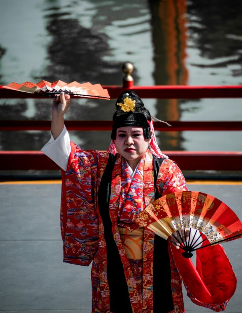 Performer in vibrant traditional Japanese attire dancing with fans near a pond