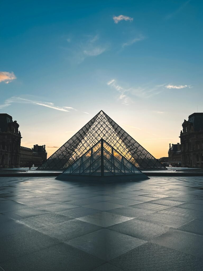The Louvre Pyramid in Paris reflecting the orange sunset sky, with the museum's classical wings behind