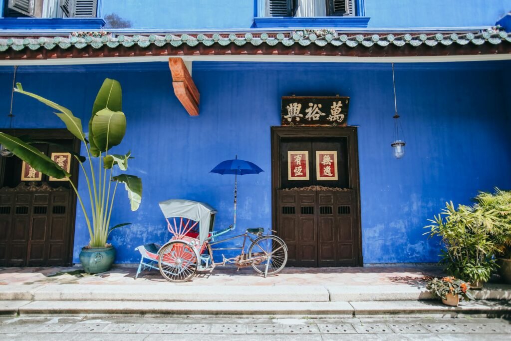 Traditional Penang trishaw parked in front of a vivid blue heritage shophouse facade in George Town's UNESCO-listed core — the iconic colonial-era streetscape that defines the city