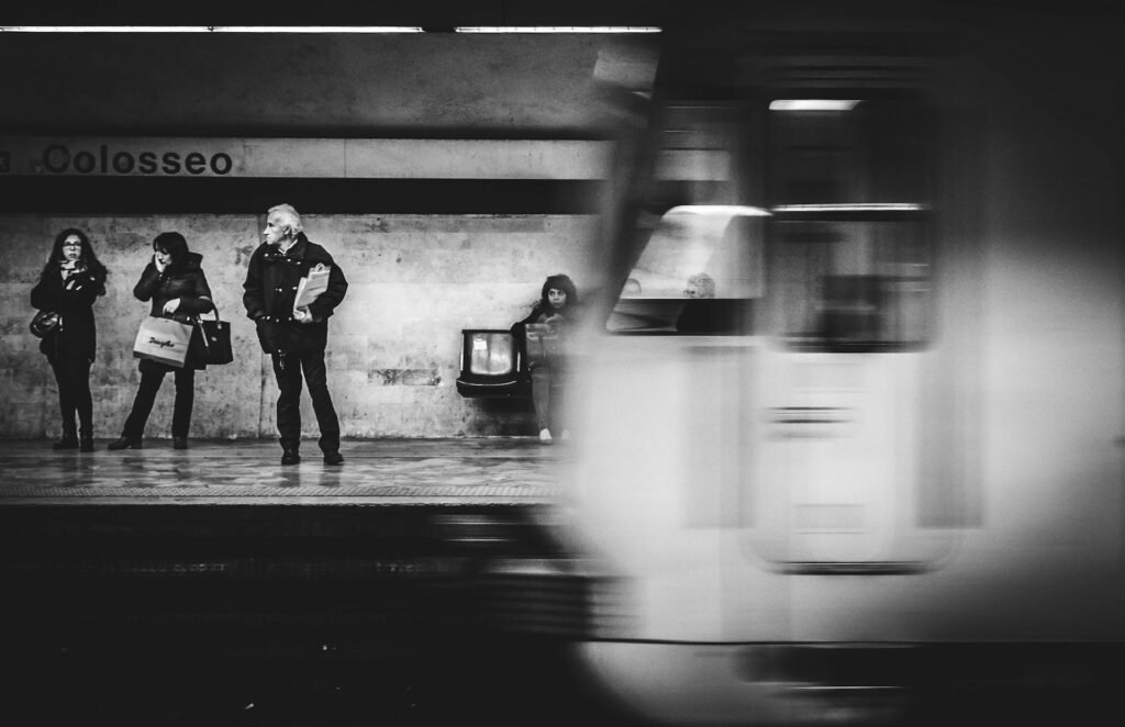 People waiting at the Colosseo Metro Station in Rome as a metro train passes by, captured in black and white