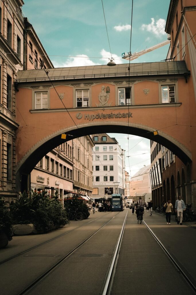 A Munich street with tram tracks passing under a historic archway, cyclists and pedestrians sharing the lane